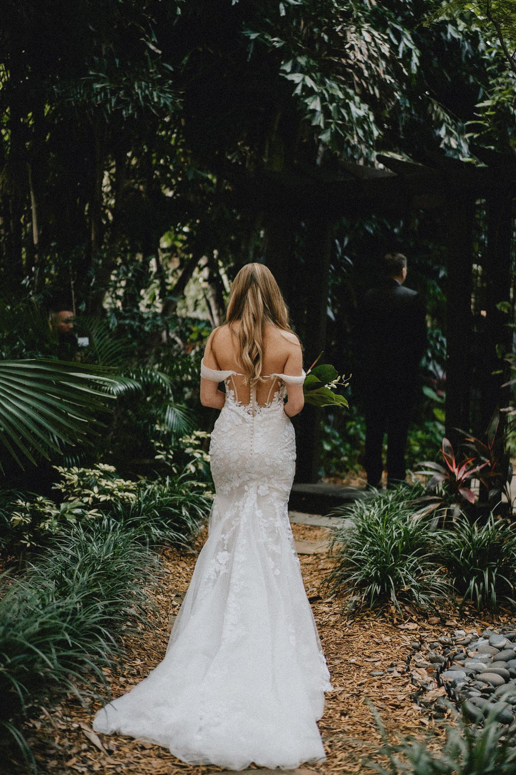 Bride in elegant lace wedding dress walking through garden path.